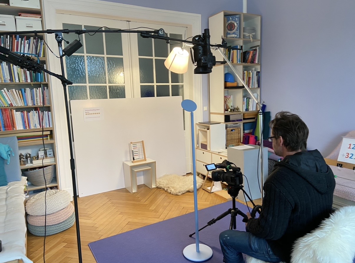 Filmszene im Montessori-Raum mit Kamera auf einem Stativ, Scheinwerfer und vorbereitetem Material auf einem Hocker.
